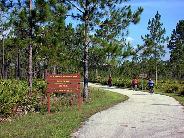 Wooden trail sign for Jay B. Starkey Wilderness Park marking the bike trail, with two cyclists riding the paved multi-use trail through pine forest in the background