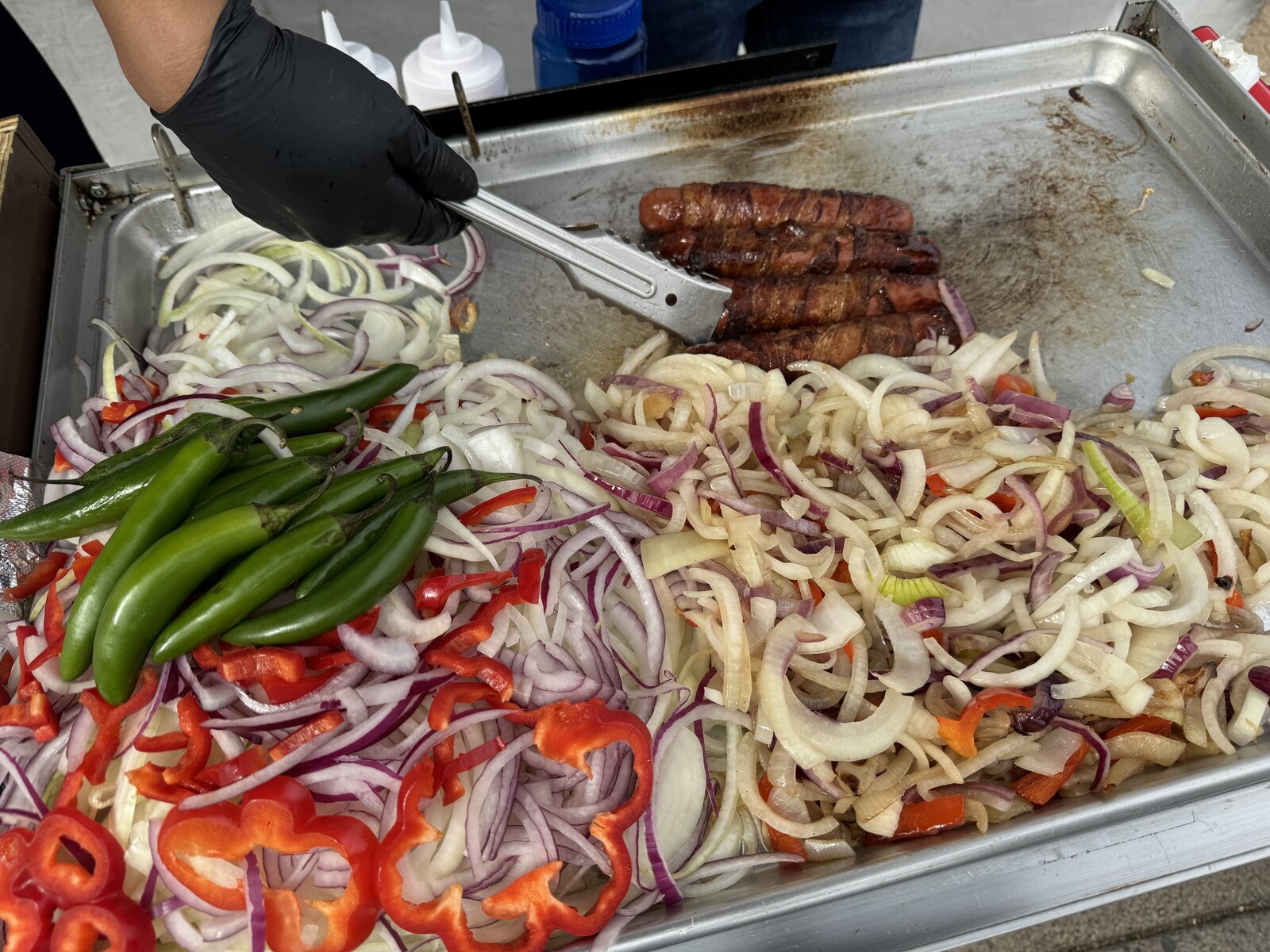 Bacon-wrapped hot dogs cooking on a griddle with onions and peppers